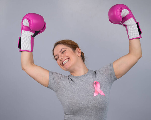 A woman with a pink ribbon on her chest raises her hands in pink boxing gloves on a gray background. Victory over breast cancer