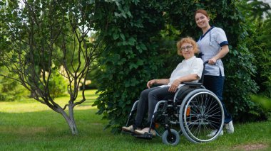 Beyaz kadın doktor yaşlı bir hastayla parkta tekerlekli sandalyede yürüyor. Hemşire dışarıda yürüyen yaşlı bir kadına eşlik ediyor.