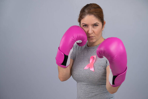 Caucasian woman in pink boxing gloves with a pink ribbon on her chest on a gray background. Fight against breast cancer