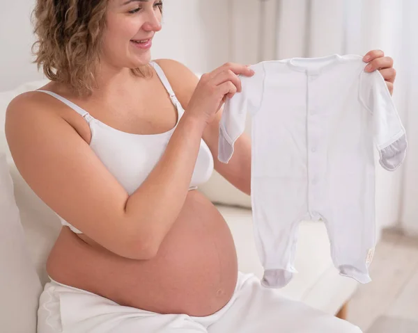 Een Zwangere Vrouw Zit Een Witte Bank Houdt Kleding Voor — Stockfoto