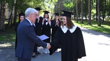 Öğretmen öğrenciyle tokalaşır ve diplomayı açık havada sunar. Bir grup üniversite mezunu.