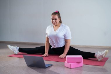 Genç, beyaz, şişman bir kadın spor minderinde viraj alıyor ve dizüstü bilgisayarında eğitim videosu izliyor. Tombul bir kız video iletişimini kullanarak ayrılığı uzaktan genişletiyor..