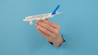 Mavi arka planda bir delikten dışarı sarkan bir kadın el bir uçak modeli tutar.