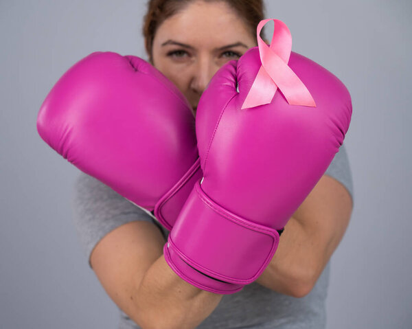 Caucasian woman in pink boxing gloves with a pink ribbon on her chest on a gray background. Fight against breast cancer