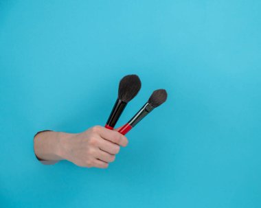 Bir kadın kozmetik için iki büyük fırça tutuyor. Kadınların elleri mavi kâğıt arkaplanından sarkıyor