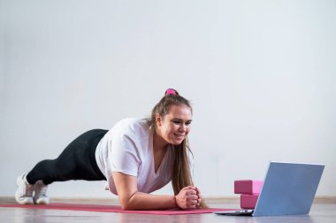 Genç, şişman, beyaz bir kadın spor paspası üzerinde tahta yapıyor. Spor kıyafetleri içinde yakışıklı bir kız spor egzersizleri yapıyor ve bir dizüstü bilgisayarda online ders izliyor..