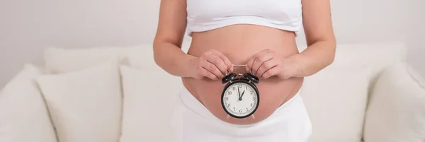 裸の腹を持つ妊婦は目覚まし時計を持っている 出生の予定日 ワイドスクリーン — ストック写真