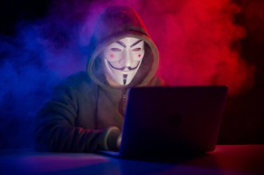 5 Haziran 2022 Novosibirsk, Rusya: Kapüşonlu Anonymous kırmızı-mavi dumanla karanlıkta dizüstü bilgisayarda yazıyor