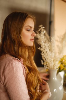 Uzun sarı saçlı, uzun pembe elbiseli, şık bir dairede, çiçekli bir kız.