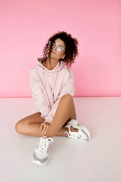 stock image Stylish young caucasian woman in light clothes in a photo studio, curly-haired woman in glasses, sneakers and hoodie, fashionable girl sitting on the floor