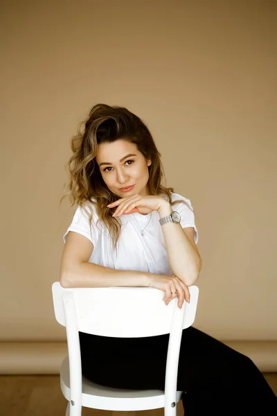 Jeune femme caucasienne en pantalon noir et un T-shirt blanc assis sur une chaise, portrait d'une brune aux yeux bruns avec maquillage et cheveux bouclés, dame en vêtements monochromes sur fond clair — Photo