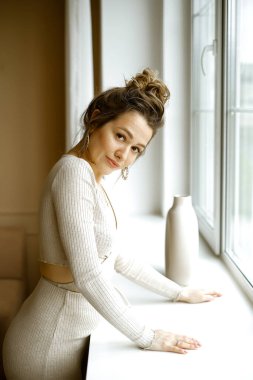 Pencerenin yanında duran bej takım elbiseli genç beyaz kadın, büyük küpeleri ve şık bir dairede yüksek saç stili olan güzel bir kız, genç ve başarılı bir kadının şık bir görüntüsü.