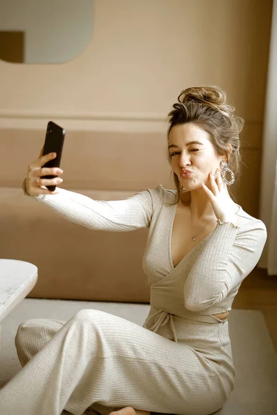 Молодая кавказская женщина в стильной квартире, смотрящая на телефон, модная дама, сидящая на полу и пишущая электронные письма, девушка-блогер в минималистской квартире с телефоном в руке — стоковое фото