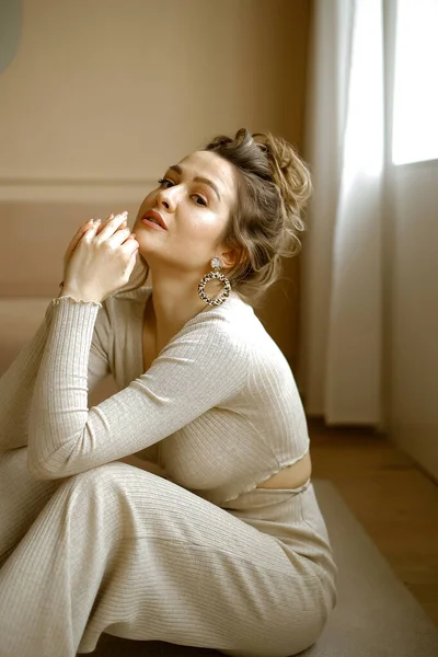 Portrait of young caucasian woman in a beige house suit sitting on the floor, a beautiful lady with large earrings and high hairstyle in a stylish apartment, a stylish image of a young successful — Stock Photo, Image