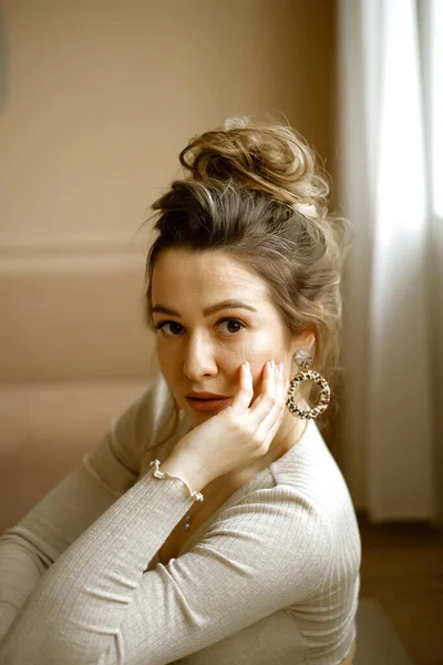 Portrait de jeune femme caucasienne dans un costume de maison beige assis sur le sol, une belle dame avec de grandes boucles d'oreilles et une coiffure élevée dans un appartement élégant, une image élégante d'un jeune succès — Photo