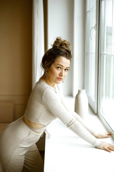 Jeune femme caucsain dans un costume de maison beige debout près de la fenêtre, une belle fille avec de grandes boucles d'oreilles et une coiffure élevée dans un appartement élégant, une image élégante d'une jeune femme réussie — Photo