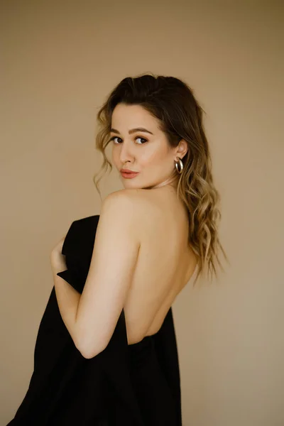 Portrait of young beauty caucasian woman with her bare back covered by a jacket , a girl with makeup and curly hair , a dark eyes lady with earrings-rings and bright makeup on light background — Stock Photo, Image