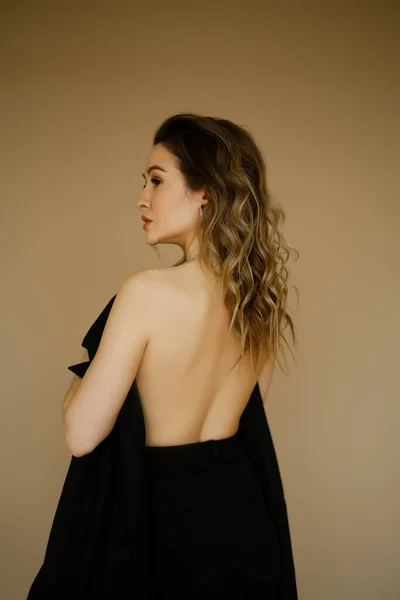 Portrait of young beauty caucasian woman with her bare back covered by a jacket , a girl with makeup and curly hair , a dark eyes lady with earrings-rings and bright makeup on light background — Stock Photo, Image