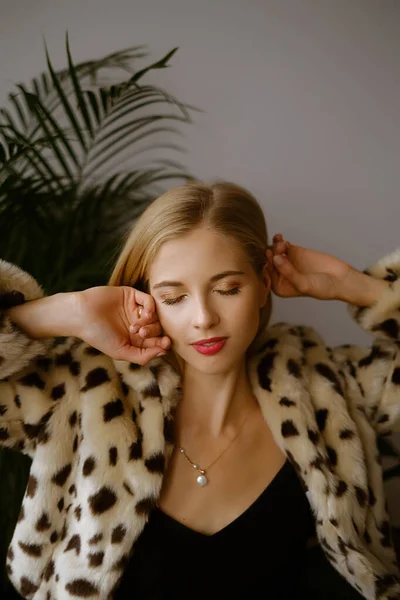 Portrait of a young caucasian woman with long blond hair wearing a leopard fur coat, the blonde closes her eyes and fixes her hair , a girl with red lips, blond hair, and a necklace around her neck — Stock Photo, Image