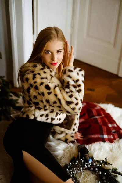 Portrait of a young caucasian woman with long blond hair wearing a leopard fur coat, a beautiful slim girl in a dress and fur coat is sitting on the floor near the Christmas tree, bright lady with — Stock Photo, Image