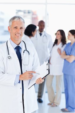 Smiling doctor pointing at a word on his clipboard clipart