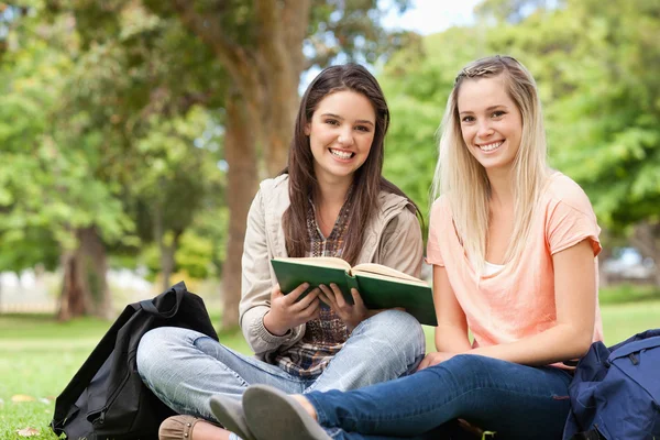 Smiling teenagers sitting while studying with a textbook — Stock Photo, Image