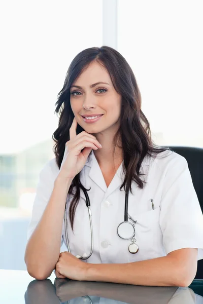 Médico sonriente sentado en el escritorio con el dedo en la mejilla —  Fotos de Stock