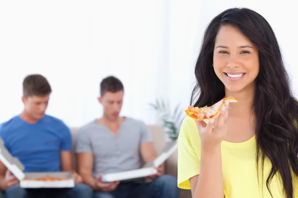 A woman holding pizza in her hand as her friends sit behind her — Stock Photo, Image
