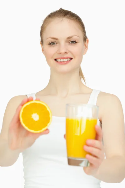 Mujer sonriente sosteniendo un vaso mientras presenta una naranja — Foto de Stock