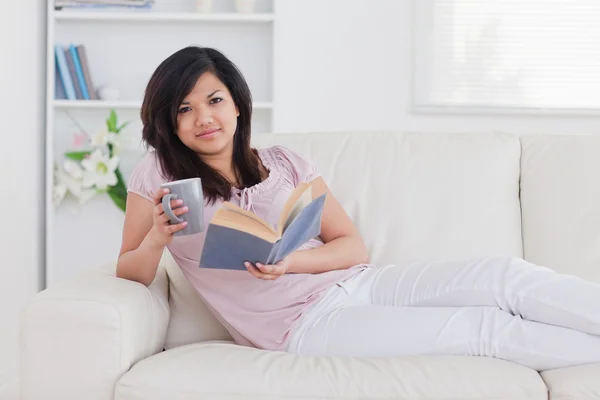 Frau liegt auf einer Couch und hält einen Becher und ein Buch in der Hand — Stockfoto