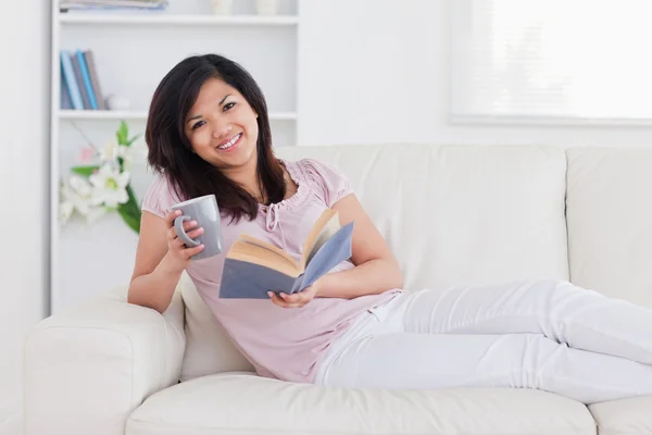 Sorrindo mulher segurando um livro e uma caneca enquanto deitado em um sofá — Fotografia de Stock