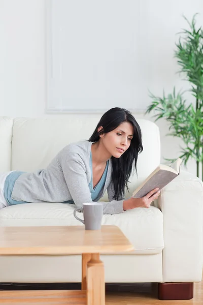 Mujer leyendo un libro con una taza en una mesa mientras descansa en un co — Foto de Stock