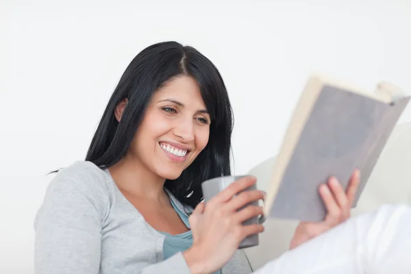 Mulher sorrindo ao ler um livro e segurando uma caneca — Fotografia de Stock