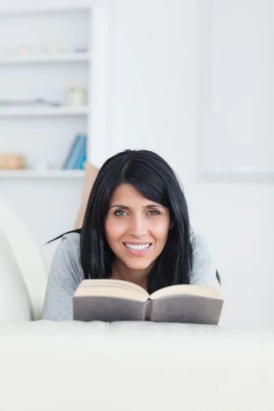 Woman holding a book while smiling and lying on a couch — Stock Photo, Image
