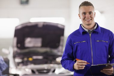 Mechanic holding a clipboard and a pen next to a car clipart