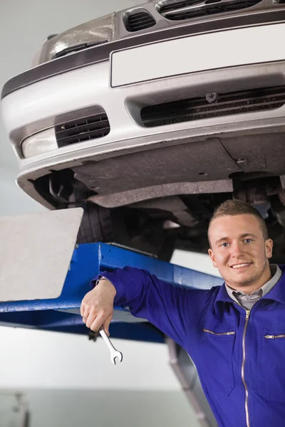 Smiling mechanic holding a spanner below a car — Stock Photo, Image