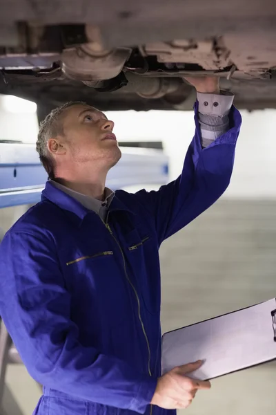 Mechanic touching the below of a car while holding a clipboard — Stock Photo, Image