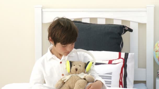 Мальчик играет с плюшевым мишкой и наушниками в постели — стоковое видео