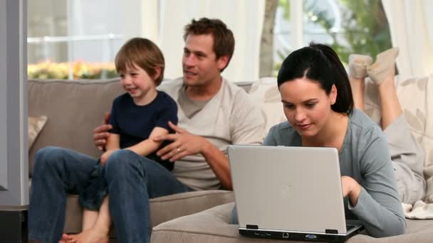 Woman using laptop while her boy and husband are watching tv — Stock Video