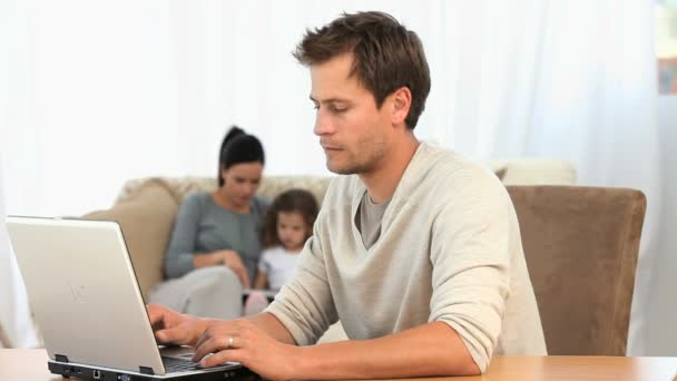 Człowiek pracy na swoim laptopie, podczas gdy jego rodzina jest na kanapie — Wideo stockowe