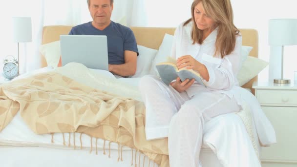 Мужчина использует ноутбук, пока его жена читает — стоковое видео