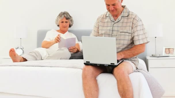 Чоловік працює на ноутбуці, поки його дружина читає — стокове відео