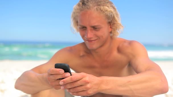 Sorrindo loiro homem sentado na areia enquanto mensagens de texto — Vídeo de Stock