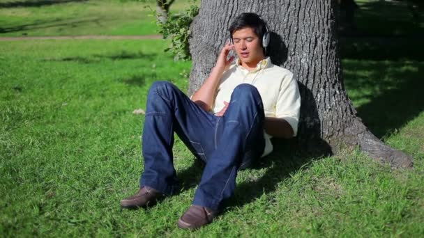 Smiling man listening to music while sitting against a tree — Stock Video