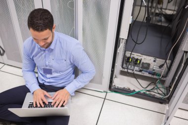 Technician typing on the laptop in front of server clipart