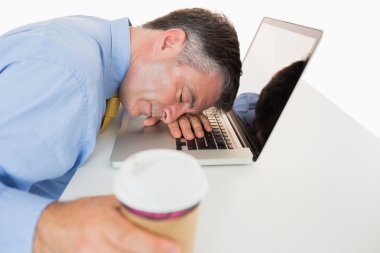Exhausted man sleeping on his laptop while holding coffee clipart