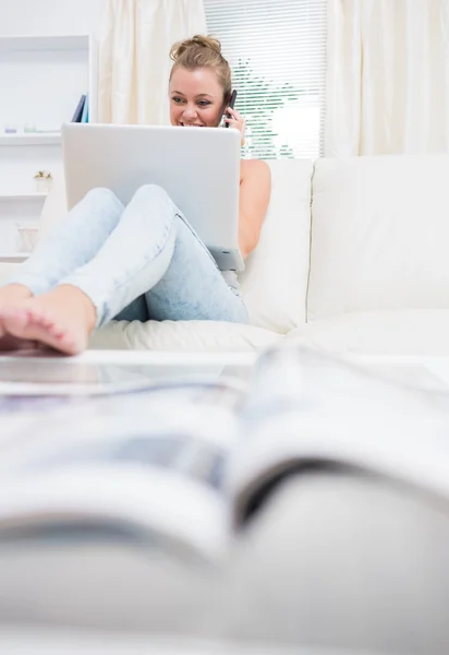 Frau sitzt auf der Couch, während sie Laptop benutzt und telefoniert — Stockfoto