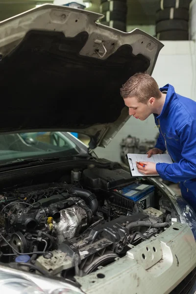 Mechanic with clipboard examining car engine — Stock Photo, Image