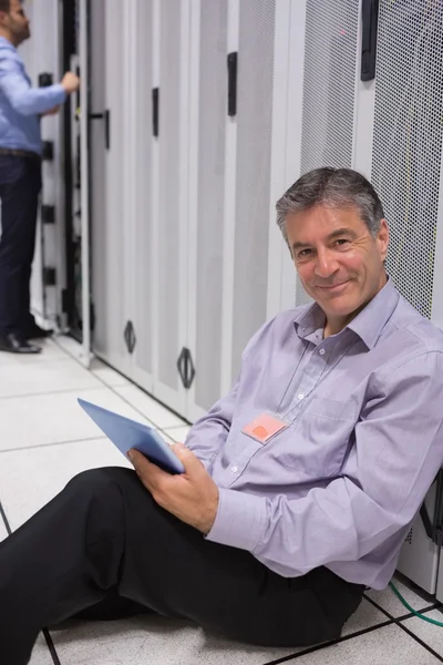 Glimlachende man zittend op de vloer met zijn tablet naast servers — Stockfoto