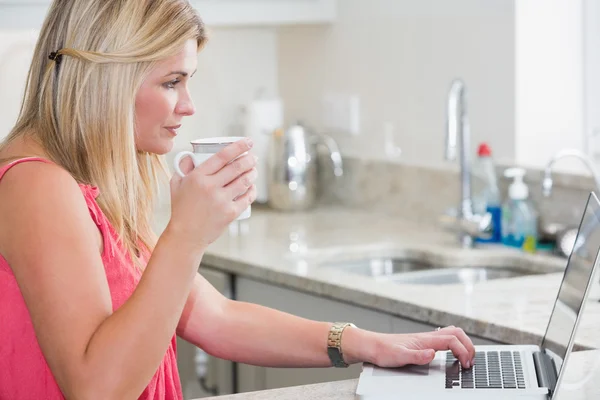 Mulher casual com xícara de café usando laptop na cozinha — Fotografia de Stock